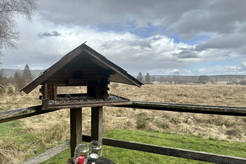 Vogelvoederhuisje op een houten terras bij La Cabane in België, uitkijkend op een open veld en bewolkte lucht.