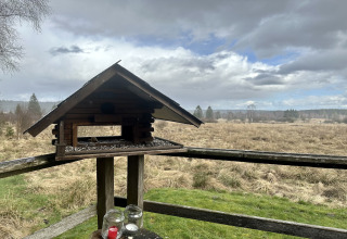 Vogelvoederhuisje op een houten terras bij La Cabane in België, uitkijkend op een open veld en bewolkte lucht.