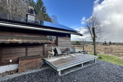 Chalet ‘La Cabane’ in België met zonnepanelen, houten terras en uitzicht op het landelijke landschap.