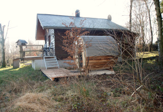Vista esterna dello chalet 'La Cabane' in Belgio con terrazza in legno, giardino e alberi intorno.