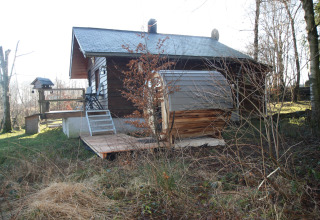 Buitenaanzicht van chalet 'La Cabane' in België met houten terras, tuin en omliggende bomen.