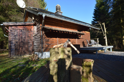 Houten chalet La Cabane in België met bijl op blok, zonnig terras en omringd door bosrijke natuur.