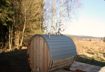 Udendørs tøndesauna ved La Cabane-chalet i Belgien, omgivet af skov og åbne marker under klar himmel.