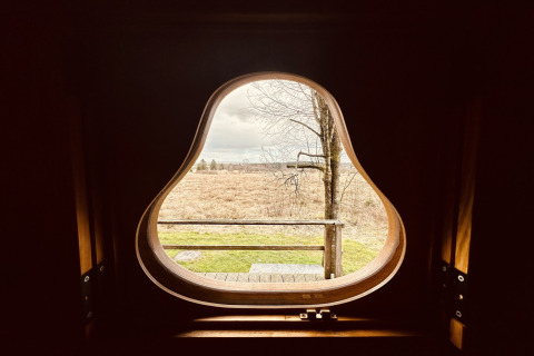 Uitzicht door een peervormig raam bij chalet La Cabane met landschap en boom in België.