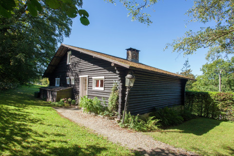 Gemütliches Blockhaus-Chalet Les Muguets in Belgien, umgeben von grüner Wiese und Bäumen bei Sonnenschein.