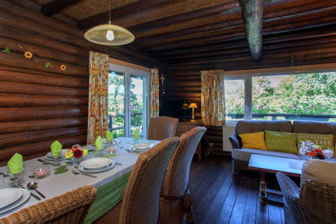 Innenraum des Chalets 'Les Muguets' in Belgien mit Esstisch, Sofa, großen Fenstern und Blumengardinen.