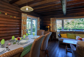 Innenraum des Chalets 'Les Muguets' in Belgien mit Esstisch, Sofa, großen Fenstern und Blumengardinen.