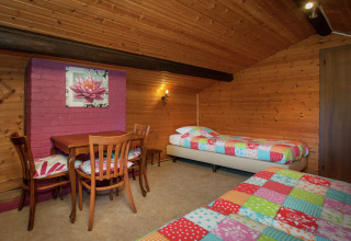 Zimmer im Chalet 'Les Muguets' in Belgien mit Holzverkleidung, bunten Betten und Esstisch mit Stühlen.