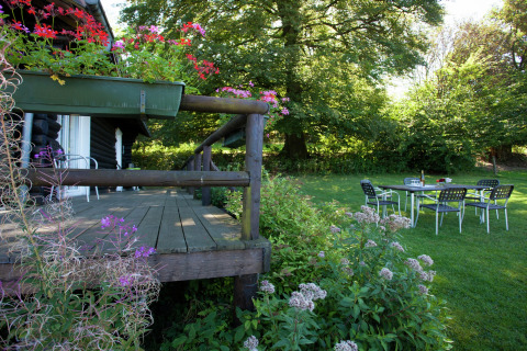 Holzterrasse mit Blumen am Chalet Les Muguets in Belgien, Gartenmöbel auf dem Rasen und Bäume im Hintergrund.