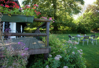 Holzterrasse mit Blumen am Chalet Les Muguets in Belgien, Gartenmöbel auf dem Rasen und Bäume im Hintergrund.