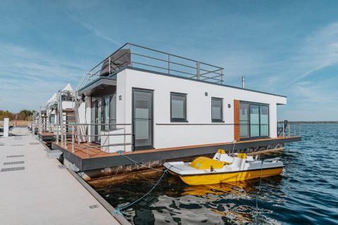 Moderne husbåd ved kajen med terrasse og gul pedalbåd, beliggende på Gräbendorfer See i solskinsvejr.