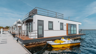 Moderne husbåd ved kajen med terrasse og gul pedalbåd, beliggende på Gräbendorfer See i solskinsvejr.