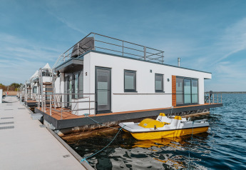 Moderne husbåd ved kajen med terrasse og gul pedalbåd, beliggende på Gräbendorfer See i solskinsvejr.