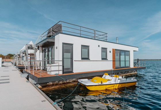 Modern houseboat docked at Gräbendorfer See, featuring terrace and yellow pedal boat on sunny day.