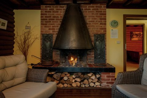 Gemütliches Wohnzimmer mit Kamin, Korbmöbeln und Holzvorrat im Chalet Les Muguets in Belgien.