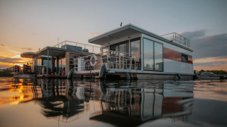 Modern houseboat in Zehdenick on calm water at sunset, showcasing reflections and a serene atmosphere.