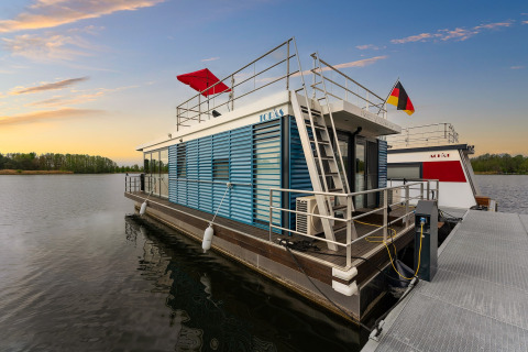 Casa flotante moderna atracada al atardecer en Zehdenick, Alemania, con vistas al agua y banderas.