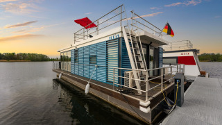 Houseboat moderna ormeggiata al tramonto a Zehdenick, Germania, con vista sull’acqua e bandiere.