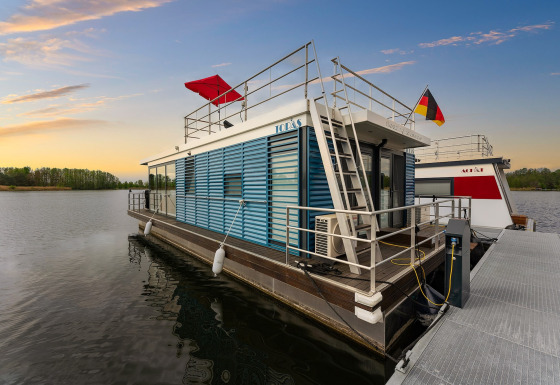 Casa flotante moderna atracada al atardecer en Zehdenick, Alemania, con vistas al agua y banderas.