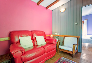 Knusse Manonfa lodge in België met rode zetel, houten stoel, kleurrijke muren en zicht op slaapkamer.