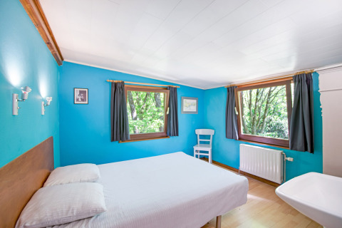 Camera luminosa al lodge Manonfa in Belgio con pareti blu, letto matrimoniale e grandi finestre sulla natura.