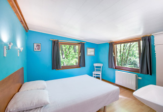 Camera luminosa al lodge Manonfa in Belgio con pareti blu, letto matrimoniale e grandi finestre sulla natura.