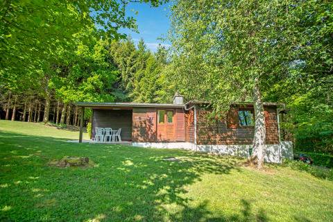 Afbeelding van het Manonfa-lodge in België, omgeven door groen gras, bomen en bos onder een blauwe lucht.