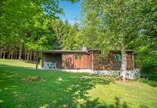 Afbeelding van het Manonfa-lodge in België, omgeven door groen gras, bomen en bos onder een blauwe lucht.