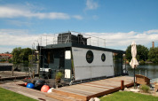 La moderna casa flotante Houseboat Friedrich atracada junto a un muelle de madera en un día soleado y claro.
