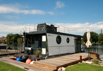Het moderne woonboot Houseboat Friedrich ligt aangemeerd bij een houten steiger op een zonnige, heldere dag.