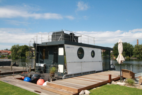 Et moderne husbåd, Houseboat Friedrich, ligger ved kajen under en blå himmel og grønne omgivelser.
