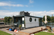 Het moderne woonboot Houseboat Friedrich ligt aangemeerd aan een steiger, omgeven door natuur en blauwe lucht.