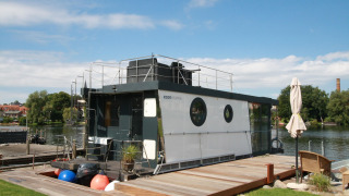 Het moderne woonboot Houseboat Friedrich ligt aangemeerd aan een steiger, omgeven door natuur en blauwe lucht.