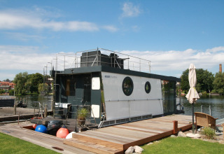Het moderne woonboot Houseboat Friedrich ligt aangemeerd aan een steiger, omgeven door natuur en blauwe lucht.
