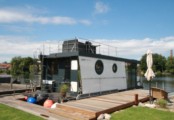 Het moderne woonboot Houseboat Friedrich ligt aangemeerd aan een steiger, omgeven door natuur en blauwe lucht.