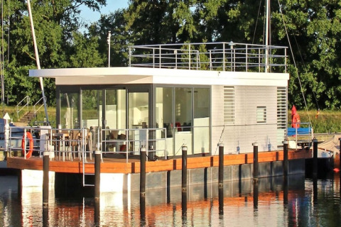 Moderne husbåd med glasfacade og tagterrasse fortøjet ved en rolig flod med grønne træer i baggrunden.