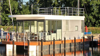 Moderne husbåd med glasfacade og tagterrasse fortøjet ved en rolig flod med grønne træer i baggrunden.