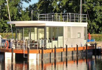 Moderne woonboot met grote ramen en dakterras aangemeerd aan een rustige oever met veel groen.