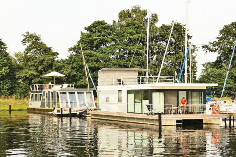 Two modern houseboats, including Houseboat Xenia, docked at a lush, green waterfront with trees behind.