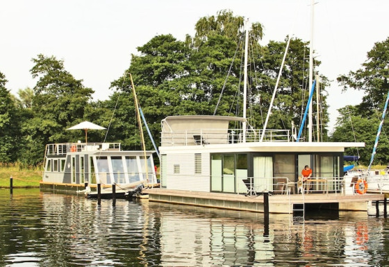 Dos modernas casas flotantes, incluyendo Houseboat Xenia, atracadas junto a un muelle arbolado y frondoso.