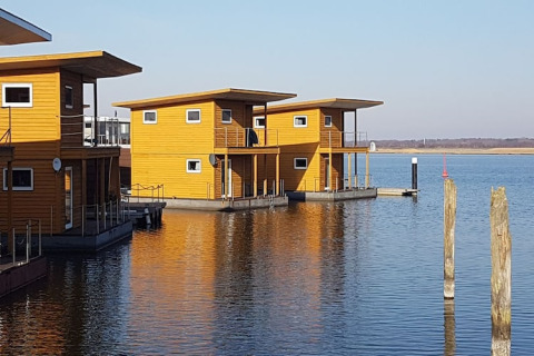Modern wooden houseboats are floating on a calm lake, with clear skies and peaceful surroundings.