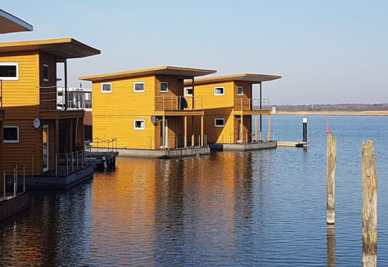 Moderne houten woonboten liggen aangemeerd op een rustige plas onder een blauwe hemel.