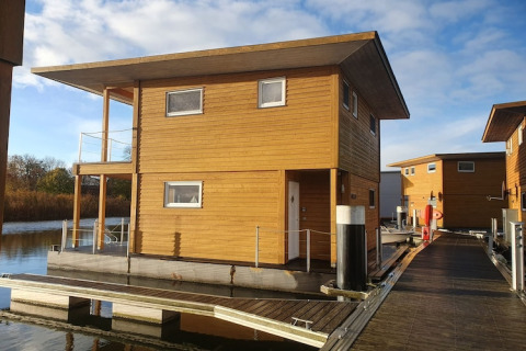 Una moderna casa galleggiante in legno è ormeggiata su un pontile galleggiante sotto un cielo azzurro.