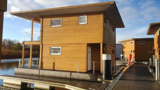 Et moderne husbåd bygget af træ ligger fortøjet ved en flydende dok under en blå himmel i solskin.