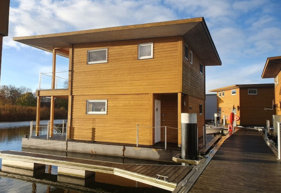 Una moderna casa galleggiante in legno è ormeggiata su un pontile galleggiante sotto un cielo azzurro.