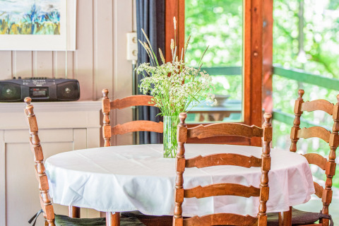 Tavolo rotondo con tovaglia bianca, sedie di legno e vaso di fiori selvatici alla lodge Manonfa in Belgio.
