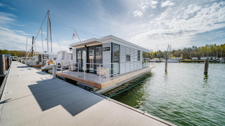 Moderne woonboot aangemeerd in Peenemünde, met zeilboten en bomen zichtbaar in de achtergrond.