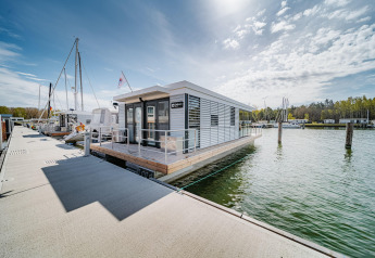 Houseboat moderna ormeggiata al molo di Peenemünde, con barche a vela e alberi sullo sfondo.