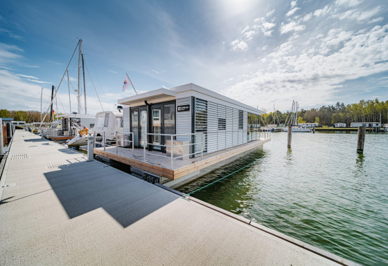 Casa flotante moderna atracada en Peenemünde, junto a un muelle, barcos y árboles al fondo bajo el cielo.