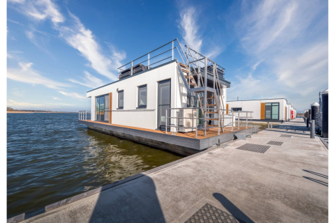 Modern houseboat in Ribnitz-Damgarten with rooftop terrace, floating on water alongside a dock in sunlight.
